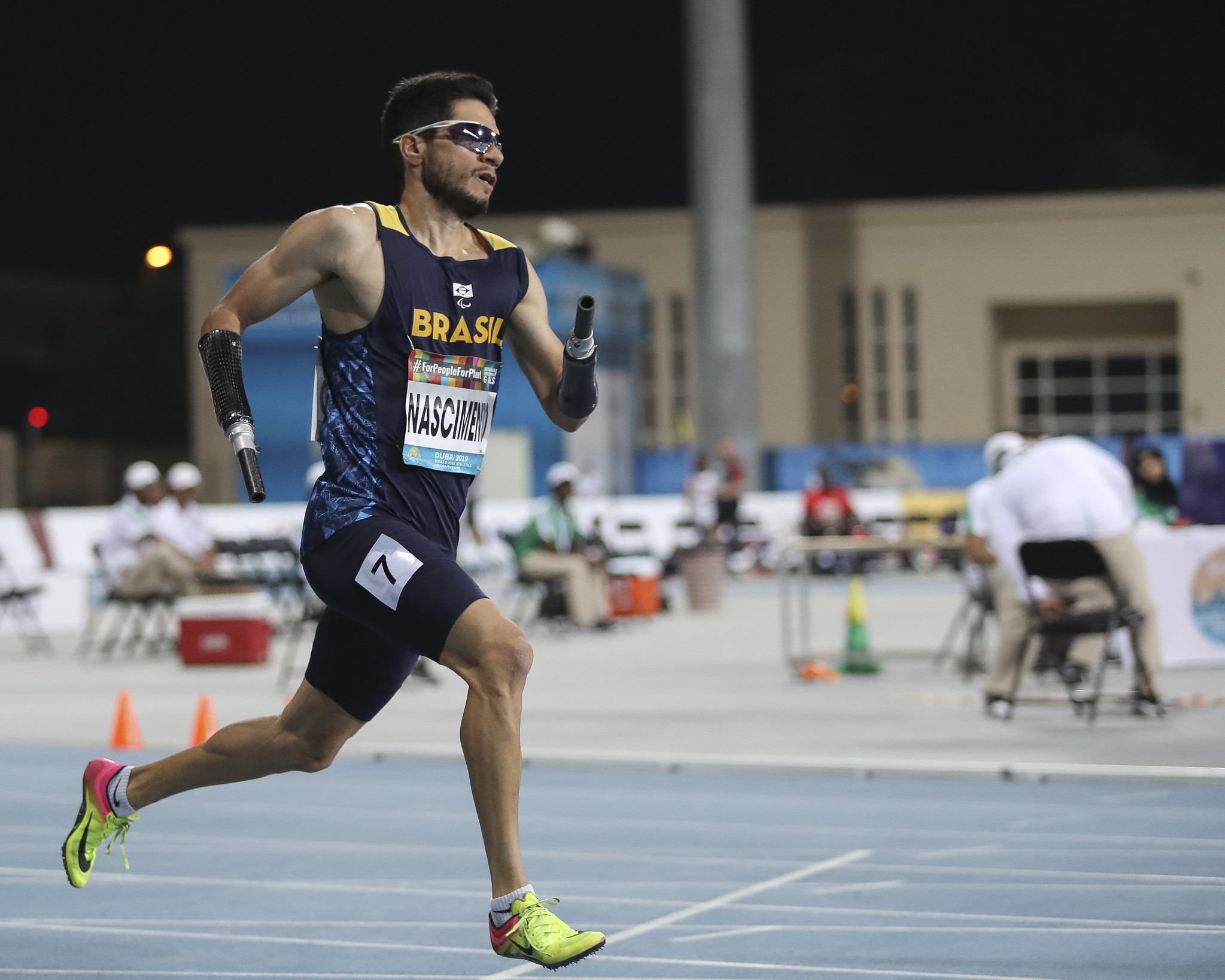 Yohansson Nascimento usa uniforme do Brasil azul escuro composto de bermuda e blusa regata, sapatilhas amarelas e óculos de sol preto. Ele corre em pista de atletismo do Mundial de Dubai utilizando próteses pretas nos braços.