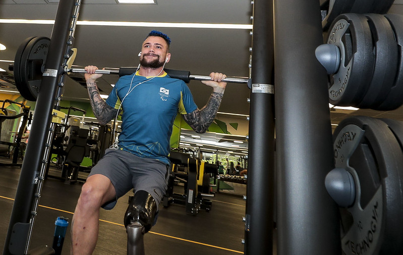 Vinícius Rodrigues, homem branco, com cabelo curto e azul, barba e braços tatuados. é amputado de perna esquerda e realiza agachamento na academia com sua prótese. Ele utiliza fones de ouvido branco, bermuda cinza e blusa azul com a logo do CPB.